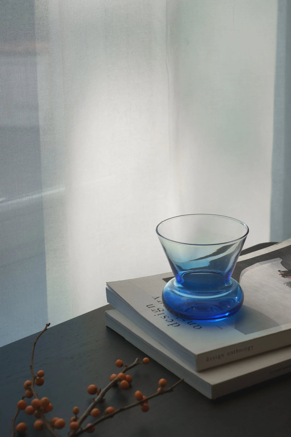 TAMO Orio Drinking Glass, a single premium blue glass with a fluted neck and wide, sturdy base, styled on stacked books. Ideal for modern home decor.