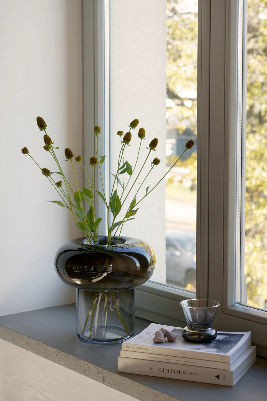 TAMO Orio Drinking Glass - Set of 2. A single dark gray, smokey glass with a unique flared top and bulbous base is displayed on a stack of books on a concrete windowsill, next to a matching vase with flowers. These contemporary luxury drinking glasses are perfect for sophisticated tablescapes.