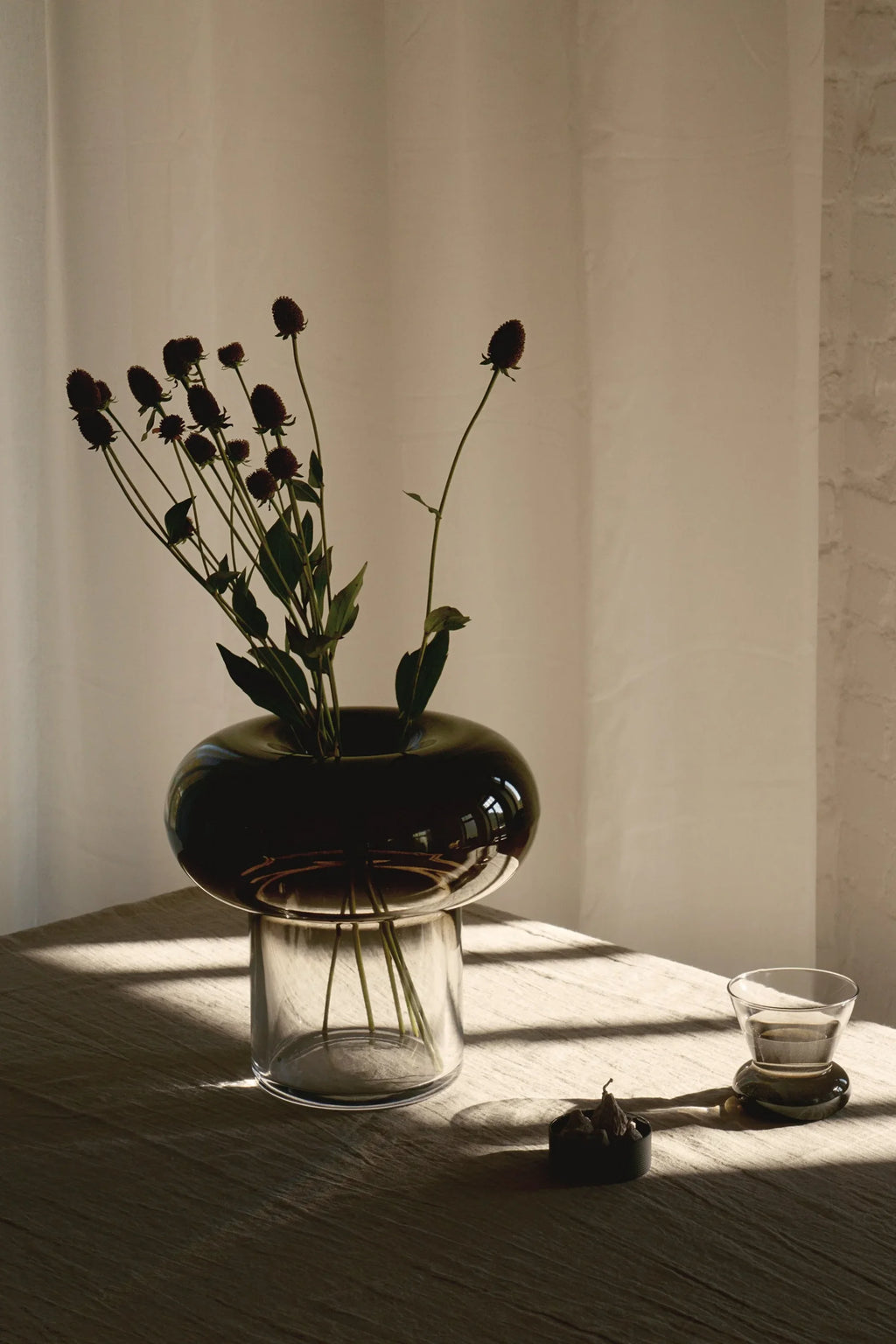 TAMO Orio Vase. Elegant mushroom-shaped dark smoke gray and clear glass vase on a lifestyle linen tabletop with dried flowers. Premium home decor accent.