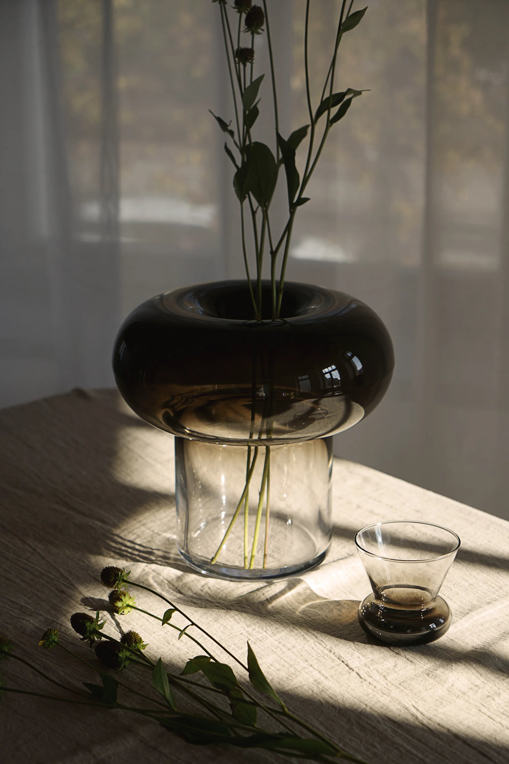 TAMO Orio Drinking Glass - Set of 2. A single smoke gray, hourglass-shaped glass, placed on a light beige linen tablecloth, bathed in sunlight. Ideal for sophisticated beverage service.