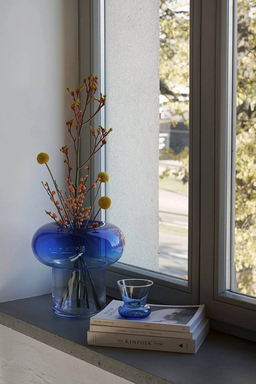 TAMO Orio Vase. A deep blue transparent glass vase, featuring a unique bulbous top sitting on a cylindrical base, filled with red-orange floral stems and yellow craspedia, displayed on a grey windowsill next to books. A luxurious, handcrafted centerpiece.