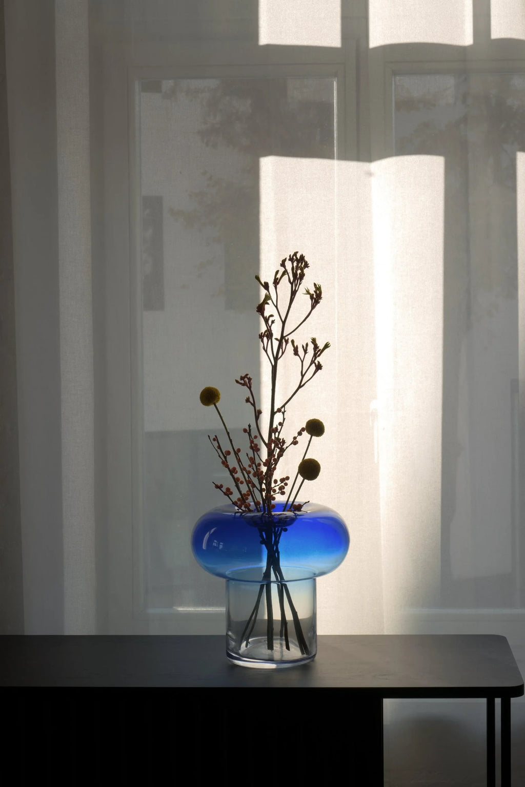 TAMO Orio Vase. Contemporary blue and clear glass vase with a unique rounded top, filled with dried, natural elements on a dark table. A stylish statement vase for any modern interior.