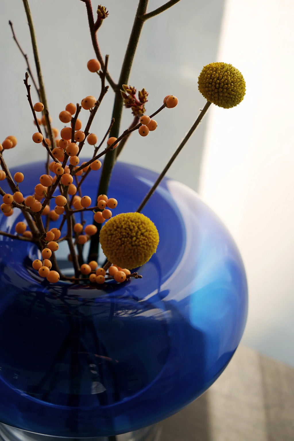 TAMO Orio Vase. Close-up of a premium, deep cobalt blue glass vase with a wide fluted neck, containing branches of vibrant orange berries and yellow spherical flowers. Perfect for elegant home decor.