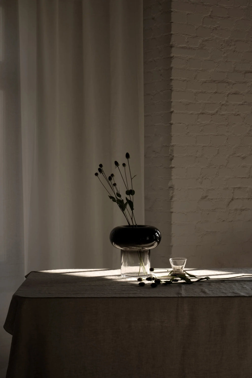 TAMO Orio Vase. A modern two-part glass vase with clear base and a dark gray, bulbous top containing dried plants, shown on a linen-covered table. An elegant, premium decorative piece.