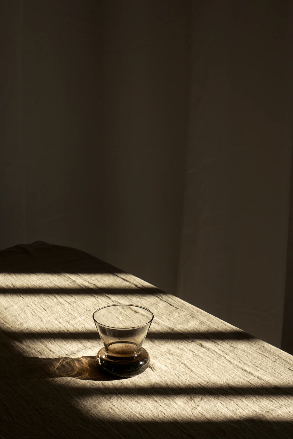 TAMO Orio Drinking Glass (Set of 2) – Single amber-toned luxury borosilicate glass with a unique bulbous base, displayed on a textured beige linen fabric with sunlit stripes. Perfect for elegant beverage serving.