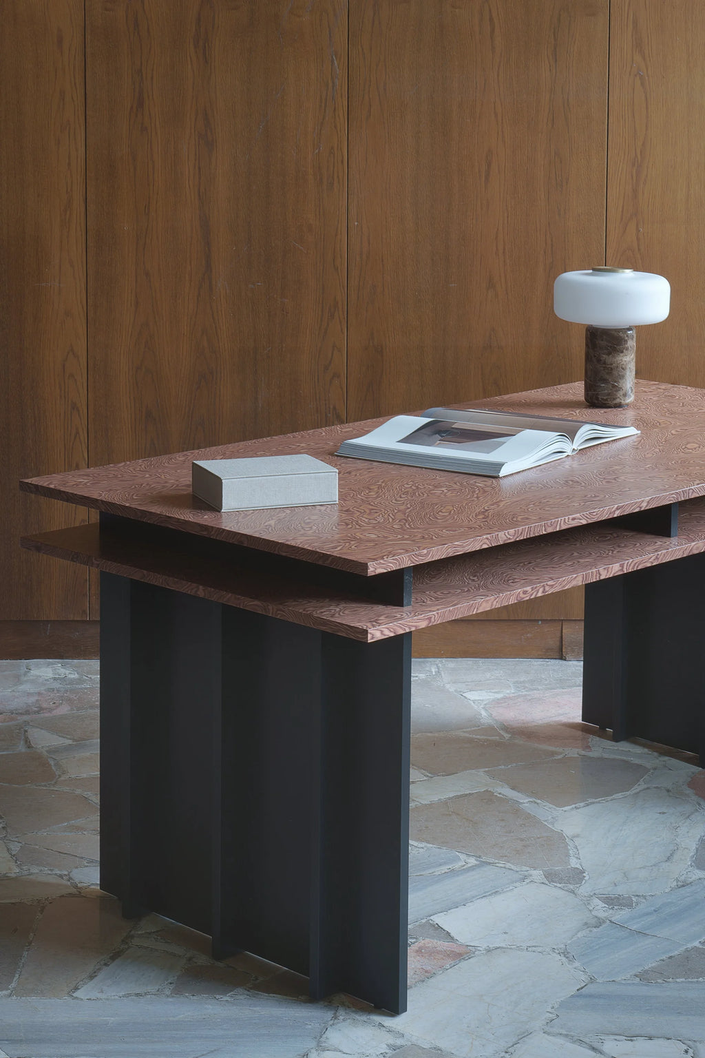 TAMO Piato Desk featuring unique reddish-brown patterned wood veneer top, black fluted legs, and a small lower shelf. This luxury desk is styled with a book, box, and white modern lamp. Ideal for a contemporary office.
