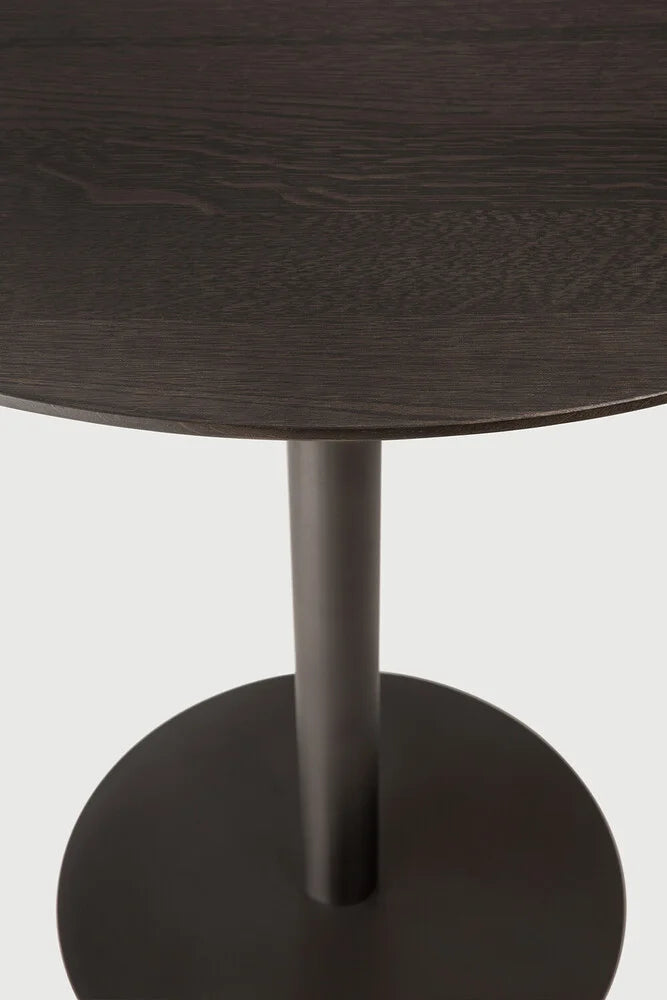 Ethnicraft Blend Round Dining Table, a modern minimalist dining table featuring a deep dark brown solid oak wood tabletop with visible grain texture, supported by a sleek matte black metal cylindrical pedestal base and round foot. Perfect as a contemporary dining room table or stylish kitchen table for modern interiors.