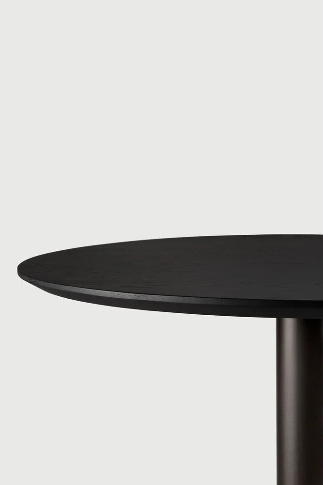 Ethnicraft Blend Round Dining Table, a contemporary minimalist dining table featuring a matte black stained solid oak round tabletop with subtle wood grain texture and a sleek, sturdy cylindrical dark metal base. Perfect as a modern dining room table or stylish kitchen table for Scandinavian and minimalist interiors.