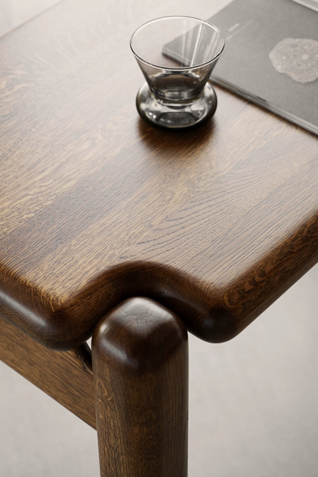 TAMO Puffo Desk, a close-up on its handcrafted dark brown oak wood desktop with visible grain patterns and elegantly rounded edges, featuring a unique integrated leg joint. A grey glass and book subtly styled on the premium wooden surface. Ideal for a sophisticated study or living room.