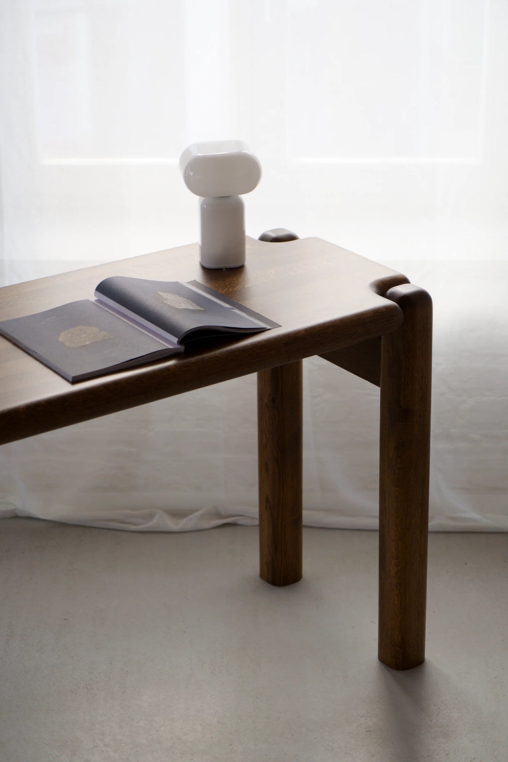 TAMO Puffo Desk, a luxury medium-brown wooden desk with rounded edges and cylindrical legs, styled with an open book and white lamp. Perfect for a chic office or living space.