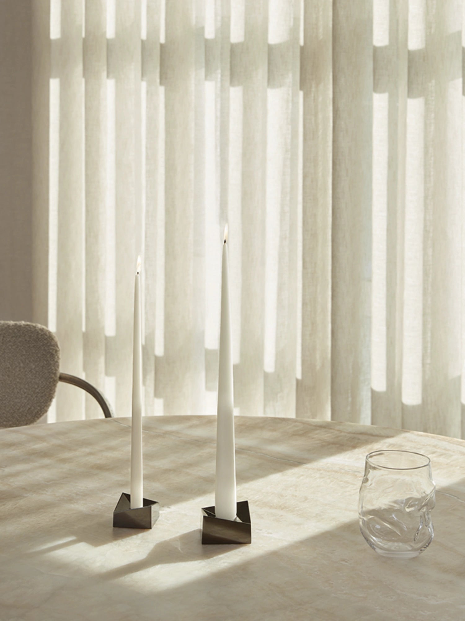 Set of 2 long-lasting and dripless taper candles in white on a beige dining table with a sculpural glass and upholstered chair.
