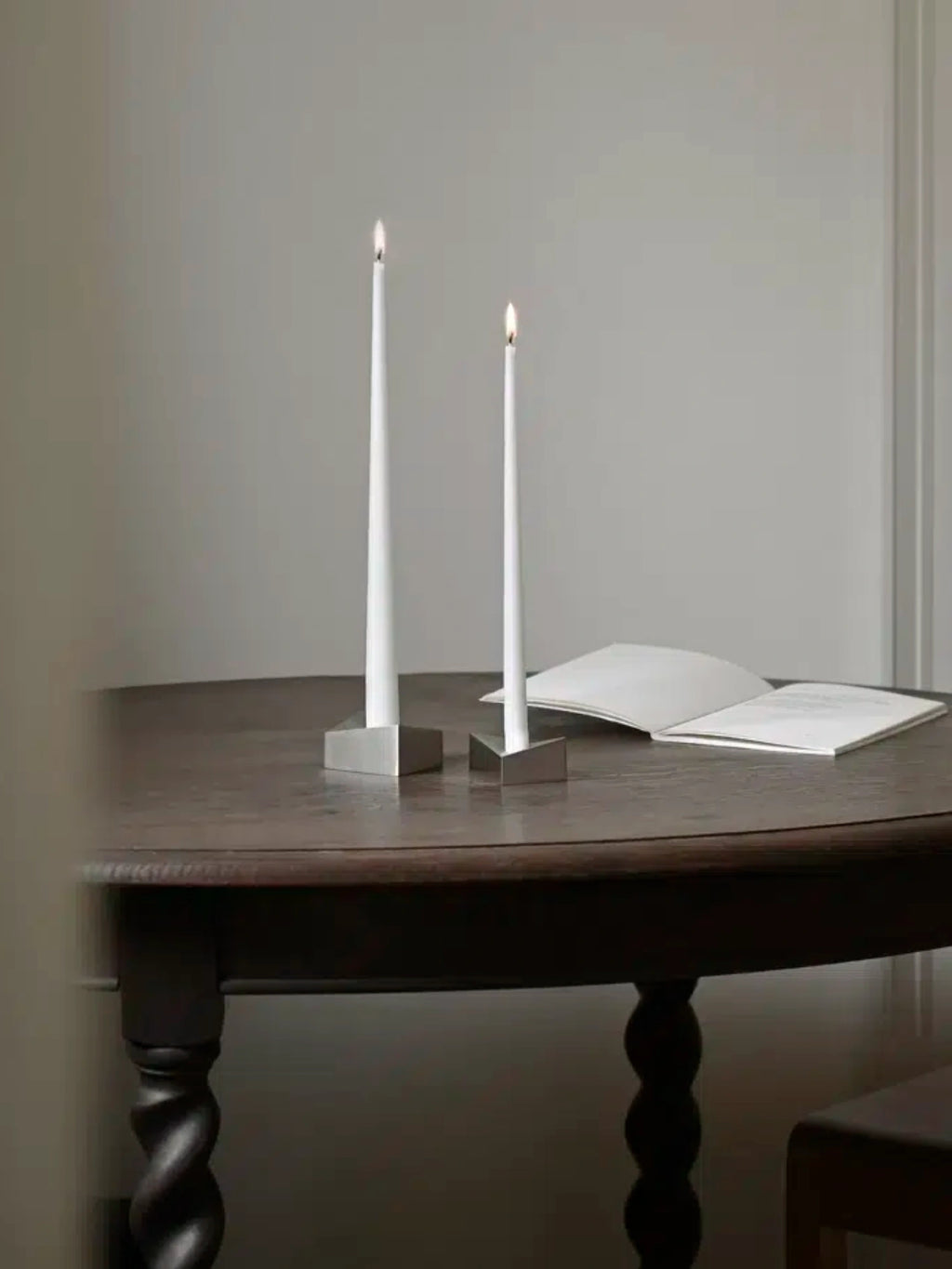 Stoff Nagel Reflect Candle Holder, a set of two modern minimalist candle holders crafted from polished silver metal with a sleek, geometric square base and a distinctive triangular cut-out designed to hold tapered white candles. The reflective surface adds a contemporary touch, enhancing ambient light and creating a striking visual element. Perfect as a sophisticated dining table centerpiece, for modern shelf styling, or as elegant Scandinavian home decor.