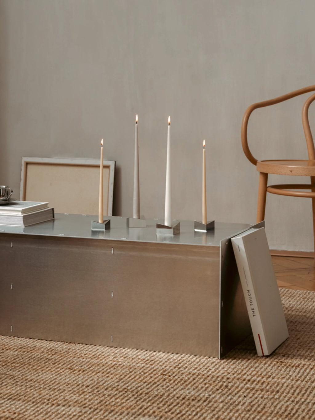 Stoff Nagel Reflect Candle Holder, a modern minimalist candle holder crafted from brushed stainless steel with a sleek, geometric triangular prism design. Its reflective silver surface holds tapering cylindrical candles in cream and light natural tones, creating a contemporary display. Perfect for industrial chic interiors, dining table centerpieces, or minimalist living room shelf styling.