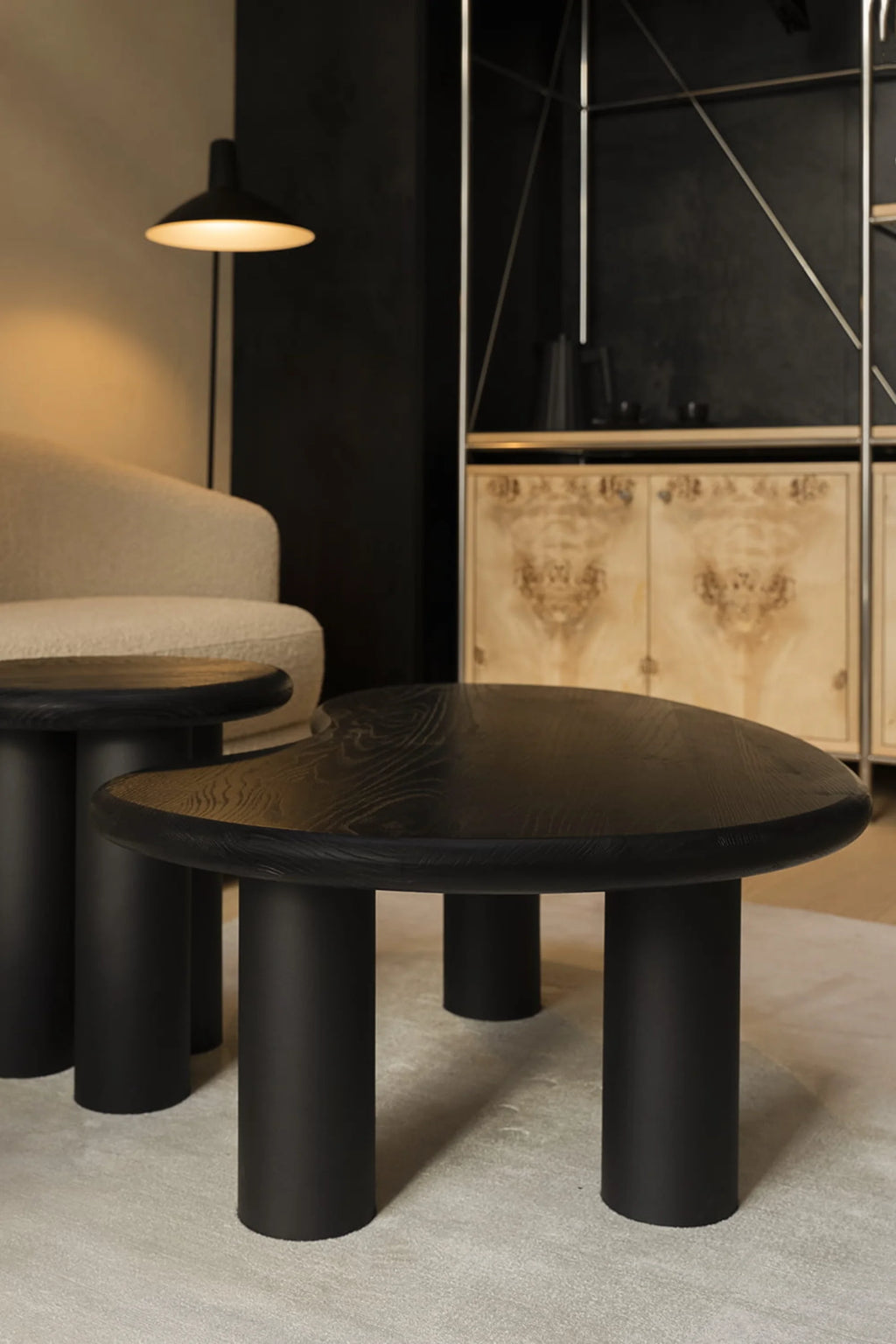 NG DESIGN Object 061 Coffee Table in a lifestyle setting, showcasing its unique, organic black ash wood tabletop with visible grain, subtly rounded edges, and four matte black cylindrical legs, beside a smaller matching table and cream textured armchair. A premium, modern centerpiece.