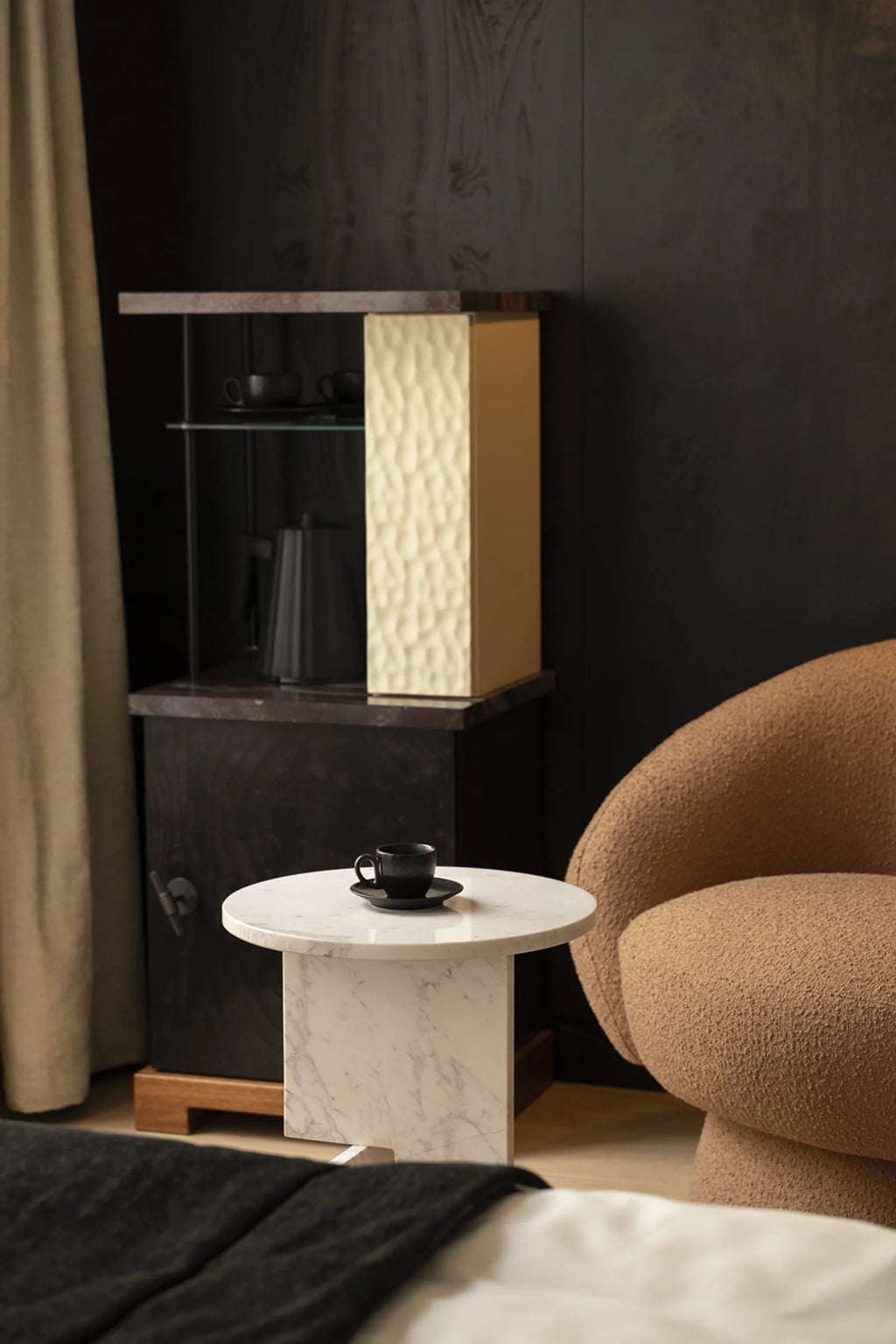 NG DESIGN Object 065 Side Table, a luxury white marble side table with natural gray veining, displayed in a sophisticated bedroom next to a textured amber armchair. A black ceramic cup and saucer rest on its round top. Ideal for a contemporary interior seeking premium accents.
