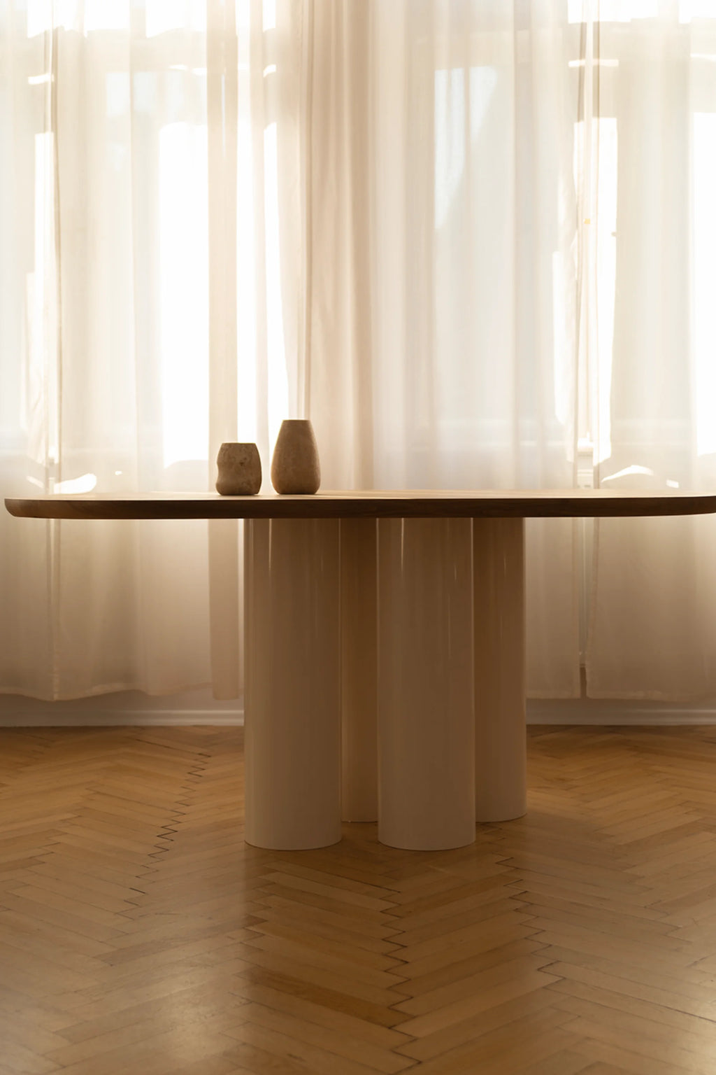 NG DESIGN Object 055 Dining Table: Oval dark wood tabletop with a light beige gloss finish and three cylindrical support legs, styled with two rustic vases on parquet flooring and sheer curtains in a sunlit room. Perfect for a modern, minimalist dining aesthetic.