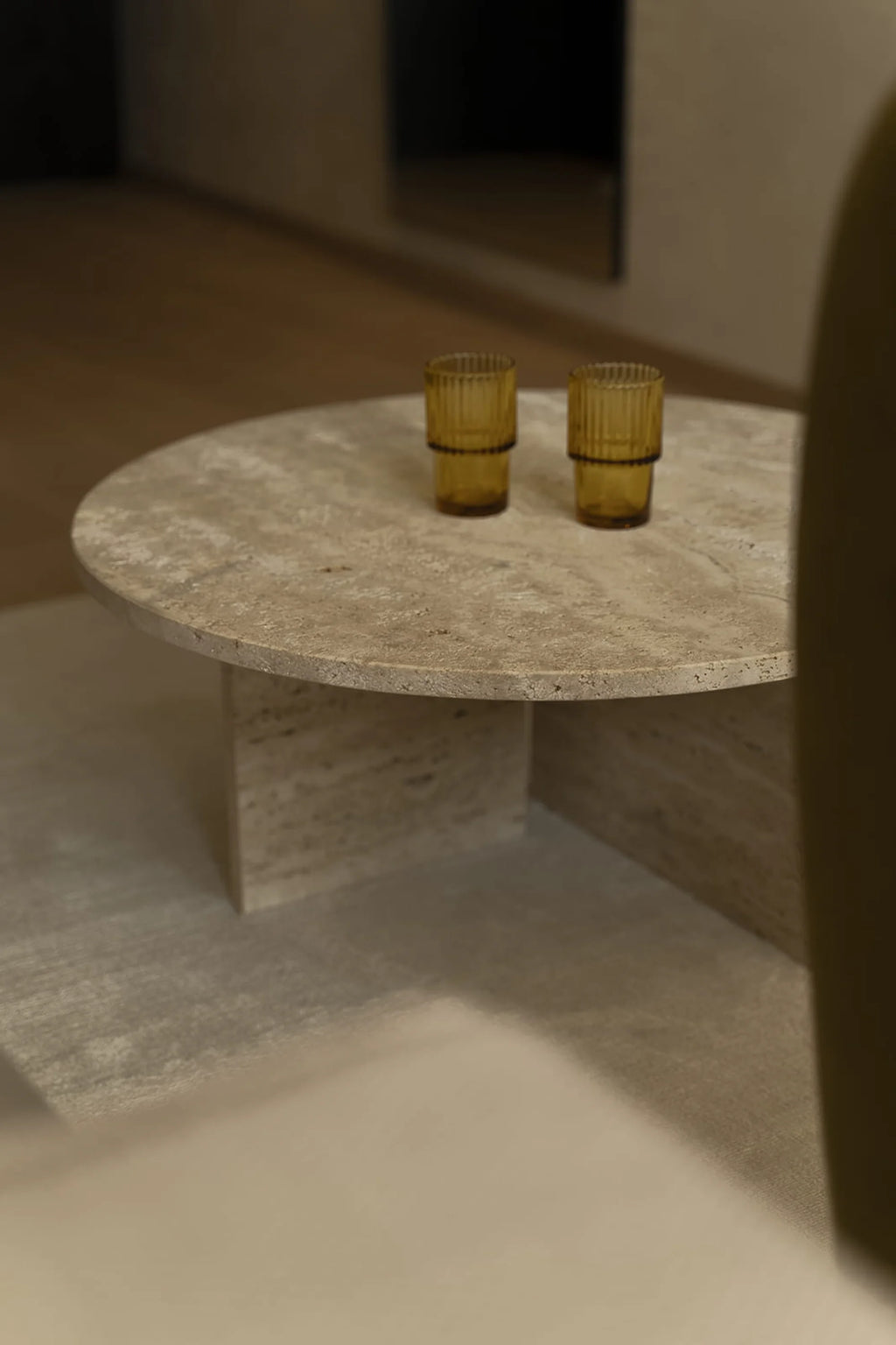 NG DESIGN Object 068 Coffee Table, a round, genuine beige/light brown travertine coffee table with a robust cross-base design, featuring two amber ribbed glasses. A luxurious centerpiece for modern living spaces.