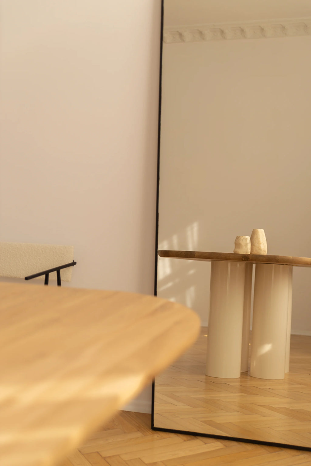 NG DESIGN Object 055 Dining Table, a luxurious light wood tabletop with a blurred foreground, and two cream-colored cylindrical pedestal bases reflected in a floor-length mirror on a warm wooden herringbone floor. Ideal for modern dining spaces.