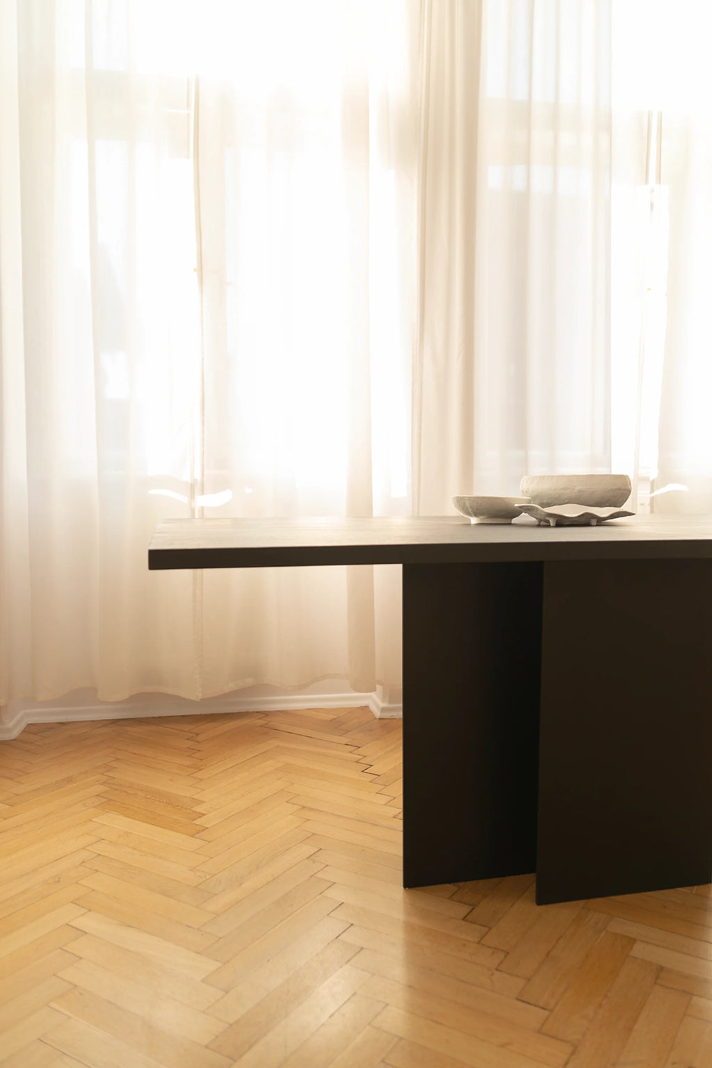 NG DESIGN Object 056 Dining Table, a minimalist dark brown wood dining table with a thick rectangular top and two flat panel legs, adorned with three textured grey ceramic bowls, showcased in a light-filled room with sheer white curtains and herringbone wood flooring. Perfect for modern living spaces.