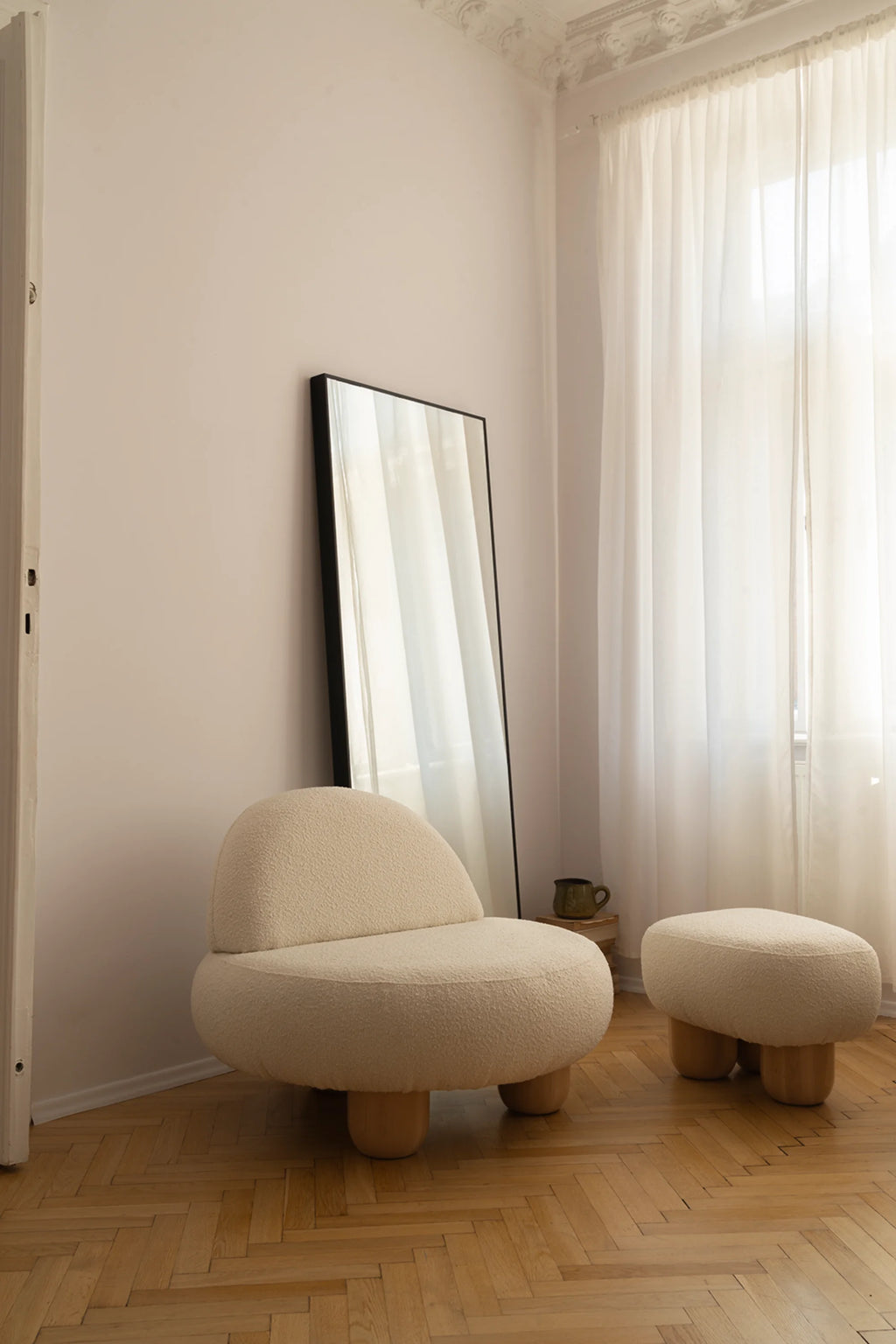 White armchair and ottoman on a wooden floor with a large mirror and curtains in the background.