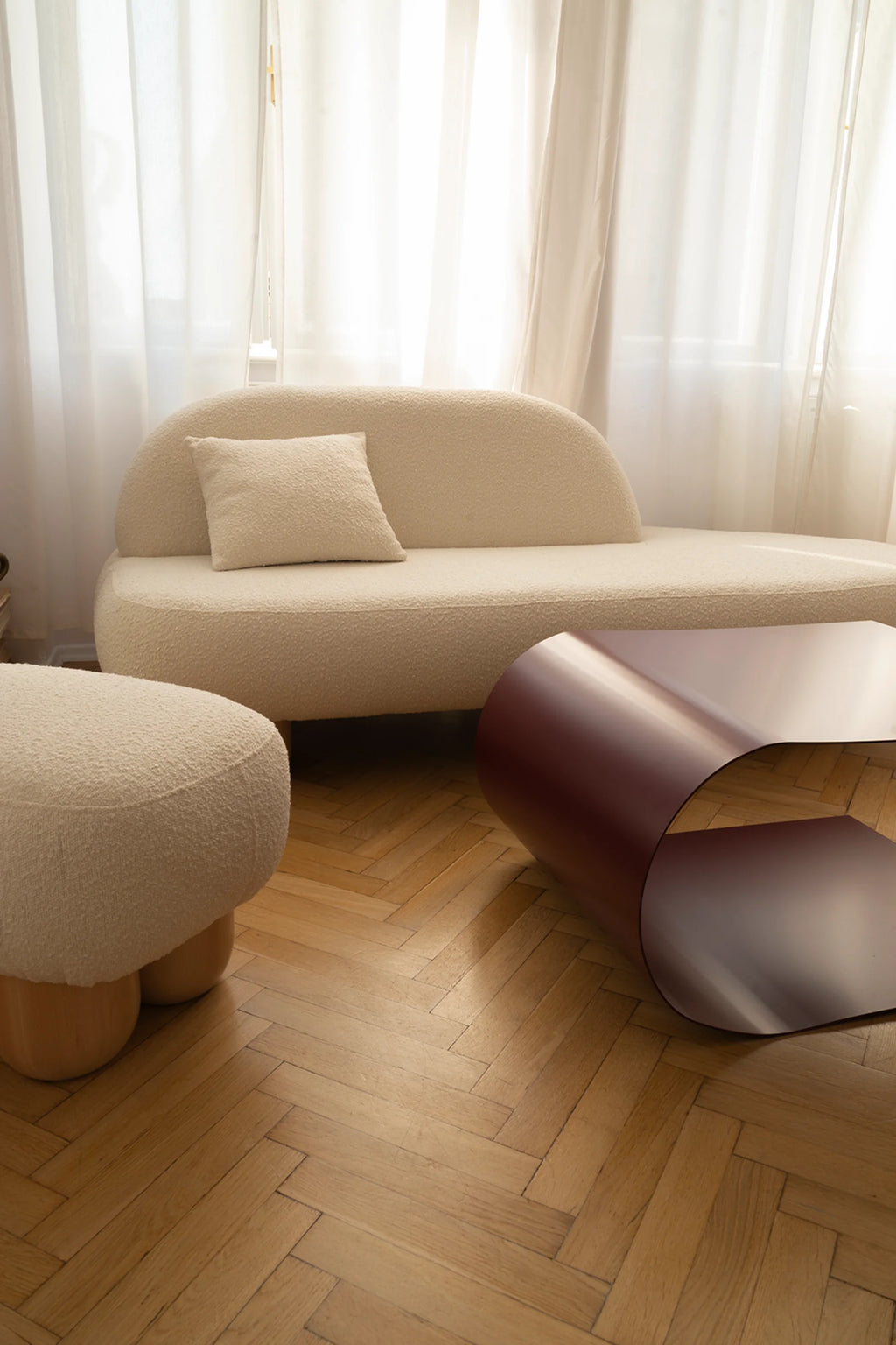 NG DESIGN Object 047 Sofa, a luxurious cream boucle curvilinear sofa with a matching throw pillow, beside a round ottoman and maroon metallic coffee table, all on a herringbone wood floor, with white sheer curtains in the background. Perfect for modern living spaces.