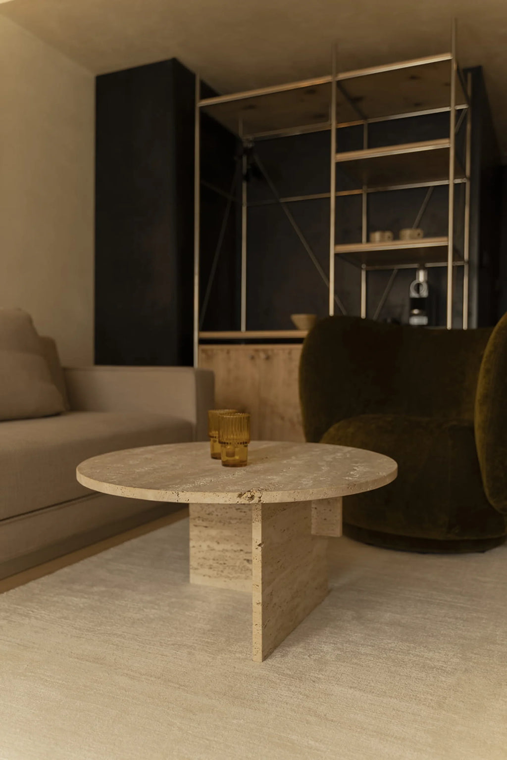 NG DESIGN Object 068 Coffee Table in a lifestyle setting, showcasing its round, thick-edged pale beige travertine top and sturdy rectangular base with visible natural porous texture. Two amber glass tumblers sit on the handcrafted table, placed on a light area rug, next to a beige sofa and dark green velvet armchair, offering a luxury centerpiece for any living space.