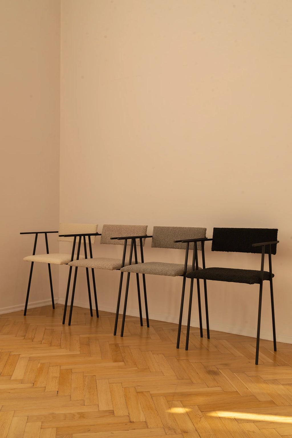 NG DESIGN Object 058 Chairs lined up; one cream, one light grey, one dark grey, and one black textured boucle fabric chair with slender black metal frames, displayed on a wooden parquet floor in a contemporary setting. Ideal for modern dining or office spaces.
