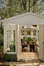 White garden shed with open doors, showing interior with plants and furniture.