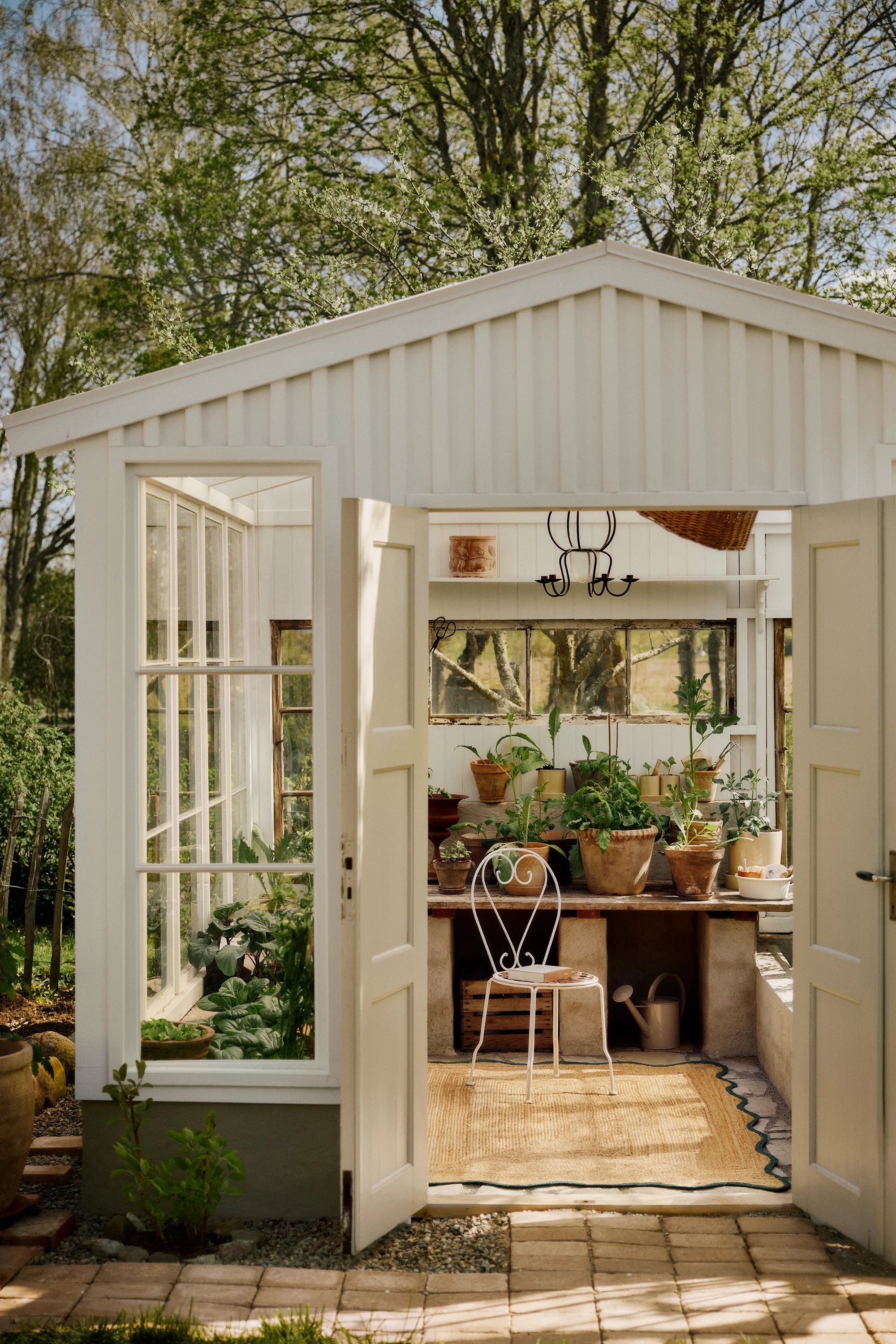 White garden shed with open doors, showing interior with plants and furniture.
