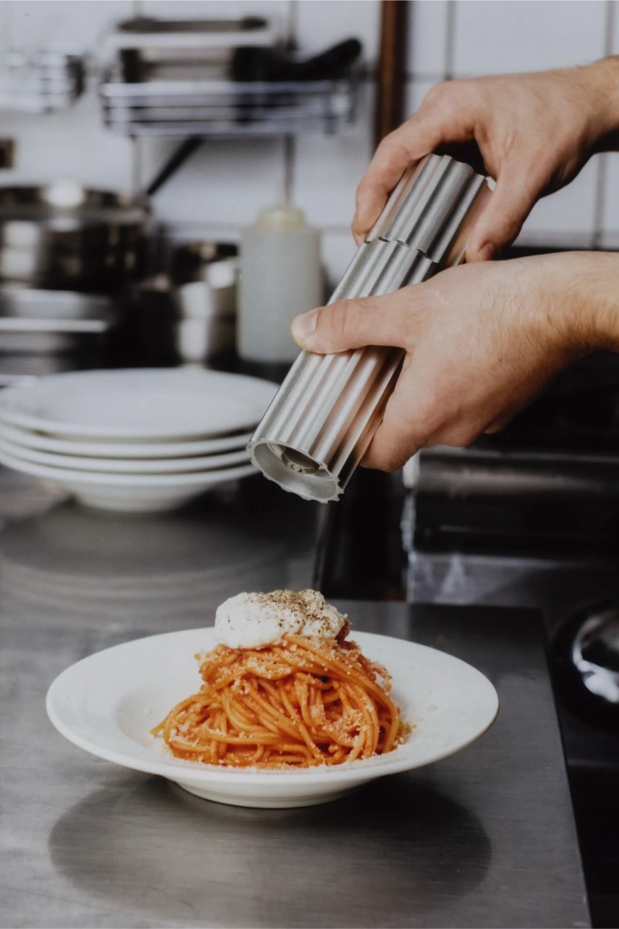 Person holding a can of pasta over a plate of spaghetti with a creamy topping in a kitchen setting.