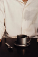 Person wearing a white shirt sitting at a table with a black coffee cup and saucer.