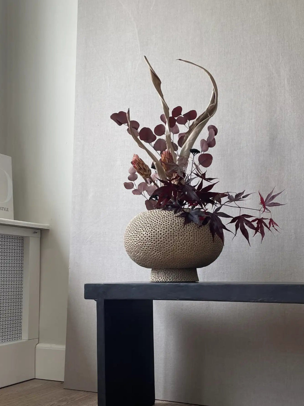 101 CPH Kabin Vase - Shisen: A handcrafted beige earthenware vase with a textured, porous, spherical body, and a smaller pedestal base, filled with dried burgundy and beige botanicals, displayed on a black console table. A statement piece for modern interior décor.