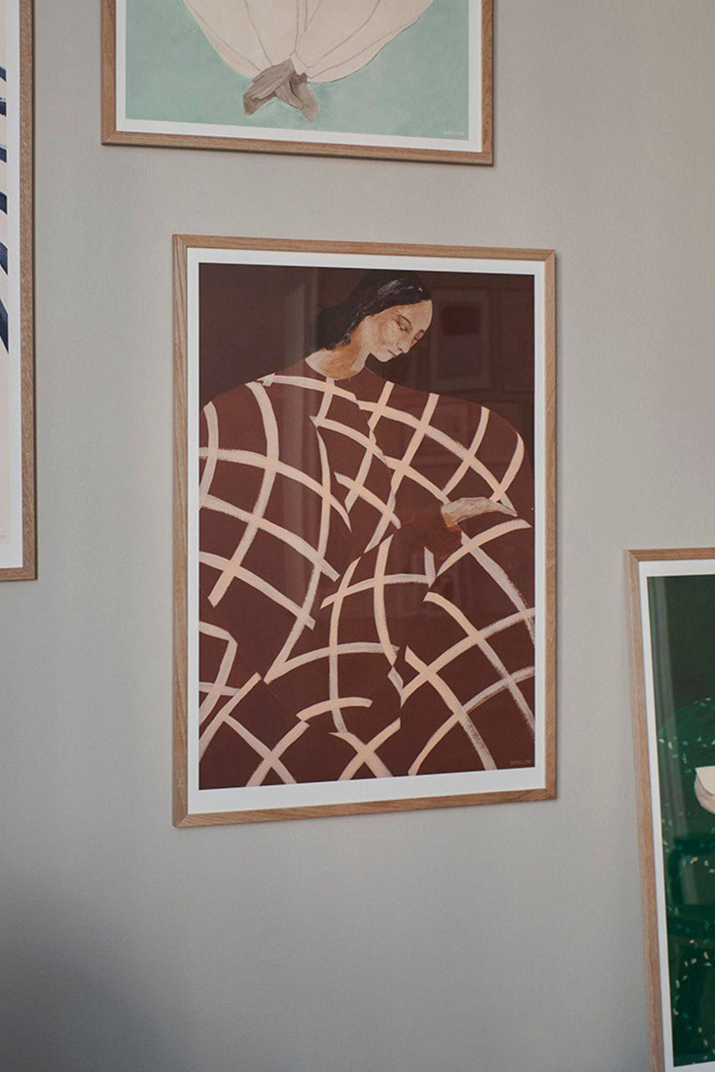 The Poster Club Waiting At Art Et Métiers, a striking contemporary abstract figurative art print depicting a woman enveloped in a sienna brown fabric with a bold pale cream grid pattern, presented in a natural oak frame with a crisp white mat. This modern wall art piece, with its earthy tones and minimalist style, is ideal for adding a sophisticated touch to Scandinavian living rooms, Japandi bedrooms, or a curated gallery wall.