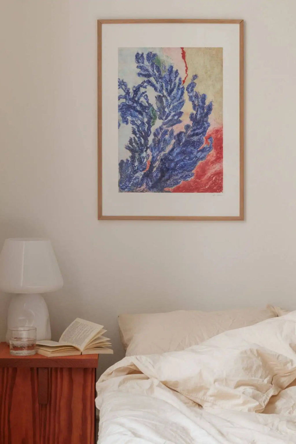 The Poster Club Quiet Bloom, an abstract art print featuring deep blue organic forms resembling coral or foliage, set against a vibrant red and muted cream background with painterly textures, framed in a natural light oak frame with a clean white mat. This contemporary wall art print is ideal for adding a serene yet bold statement to modern bedrooms, Scandinavian living rooms, or curated gallery walls.
