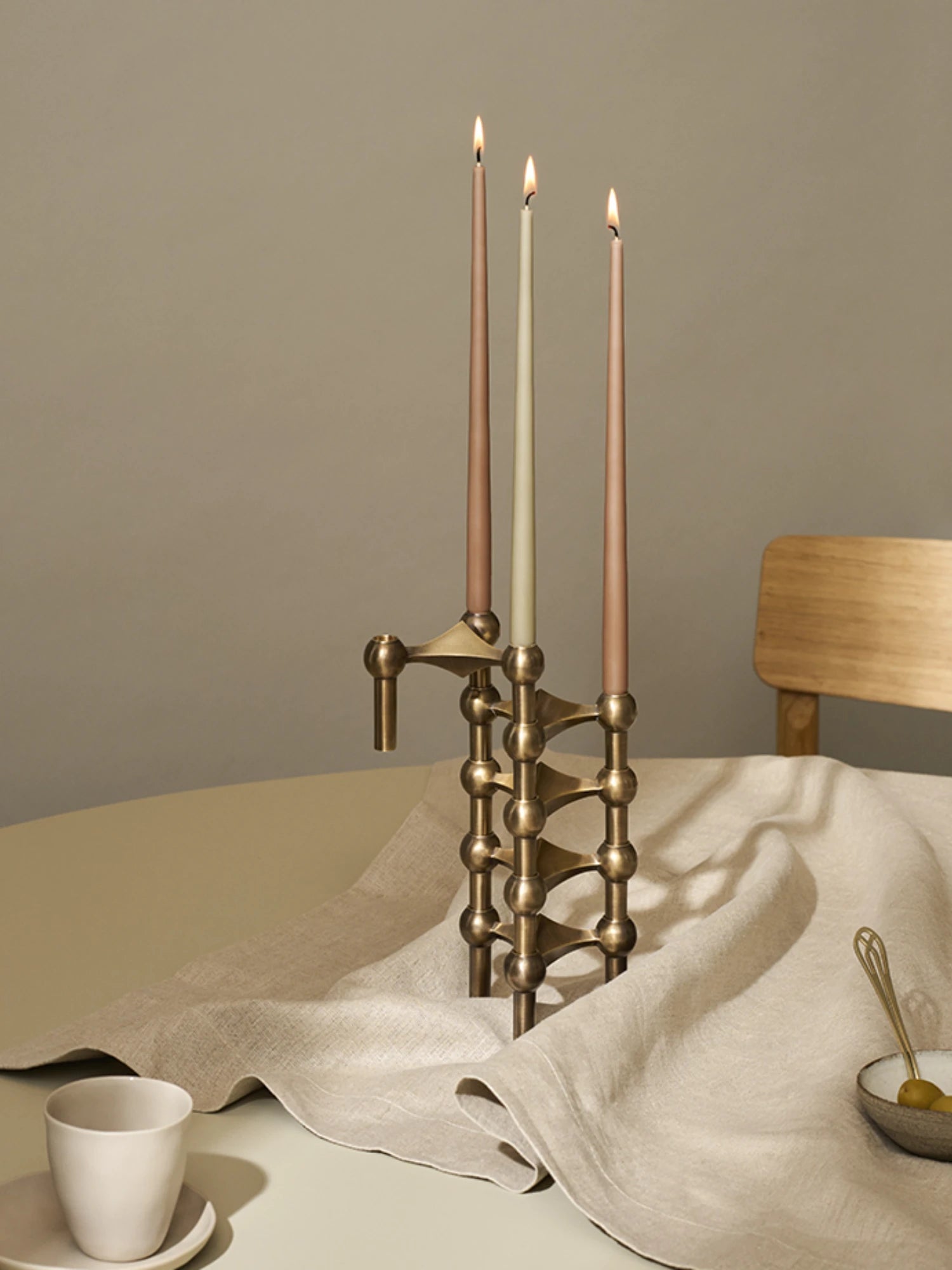 Bronze stoff nagel candle holder with three lit mix color taper candles on a dining table with a beige cloth and wooden chair.