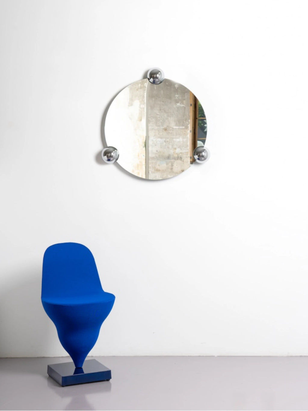 Moustache Sphera Orbit Mirror, a contemporary circular mirror with three shiny chrome spheres, reflecting a concrete wall texture, positioned above a stylish blue chair on a light grey floor.