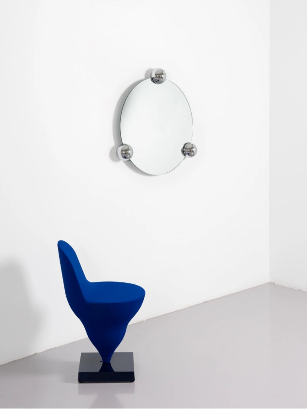 Moustache Sphera Orbit Mirror, a minimalist round mirror featuring three polished chrome spherical accents, mounted on a clean white wall above a striking blue modern chair with a unique base.