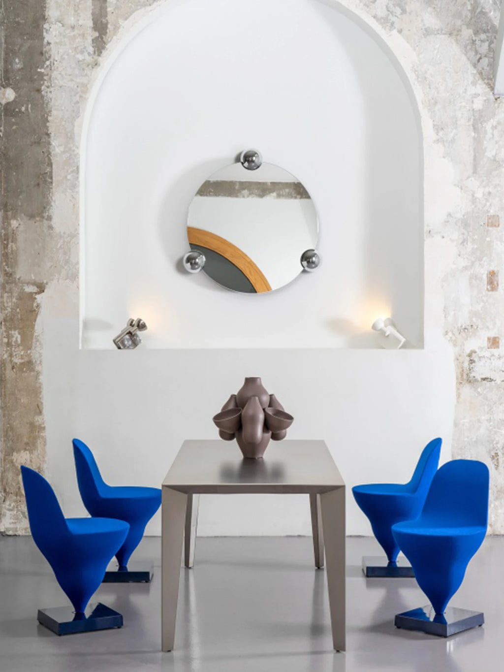 Moustache Sphera Orbit Mirror, a modern round mirror with a unique wooden accent and three polished chrome spheres, elegantly displayed in a white arched alcove above a dining table with blue chairs.