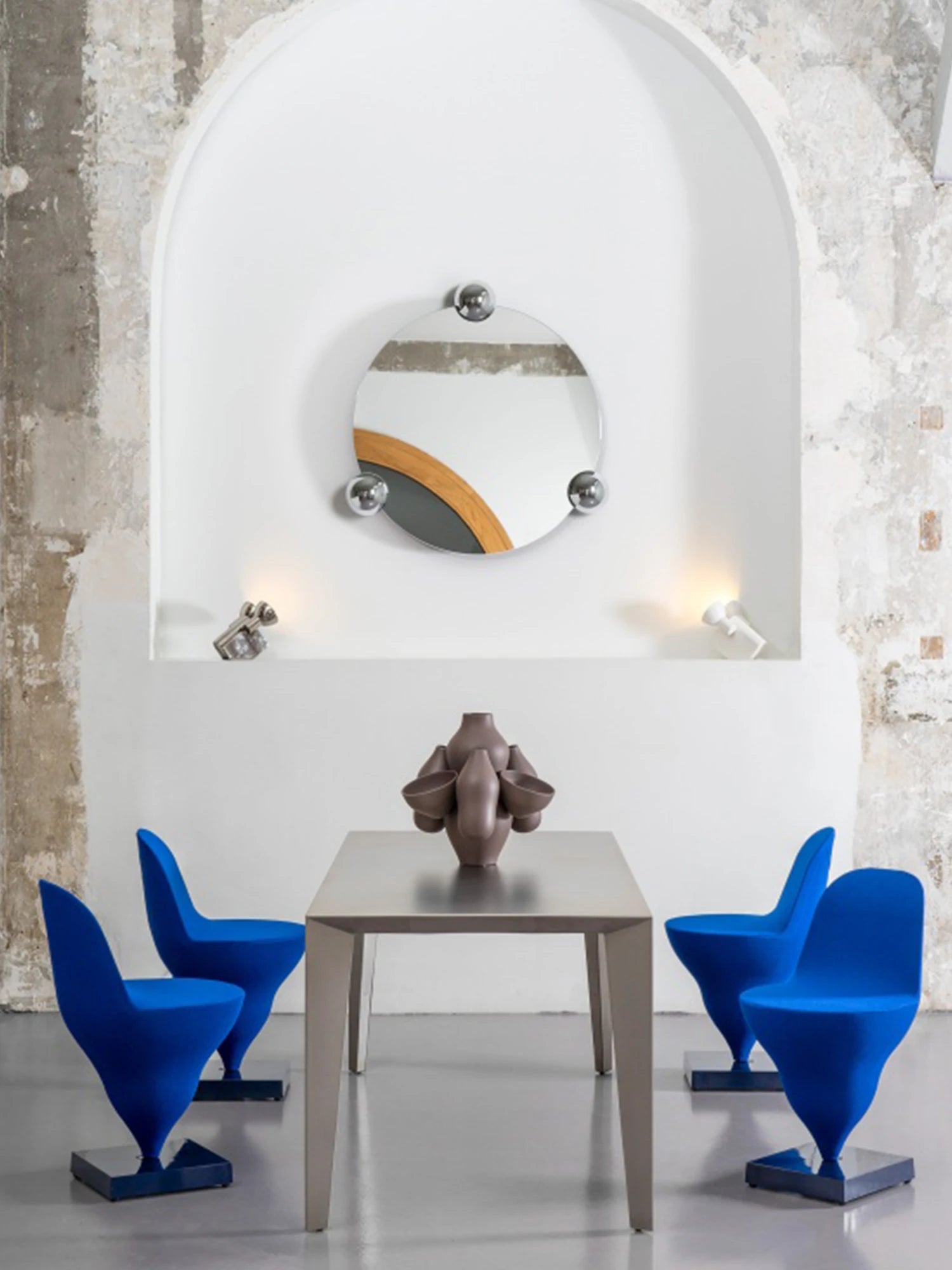 Moustache Sphera Orbit Mirror, a modern round mirror with a unique wooden accent and three polished chrome spheres, elegantly displayed in a white arched alcove above a dining table with blue chairs.