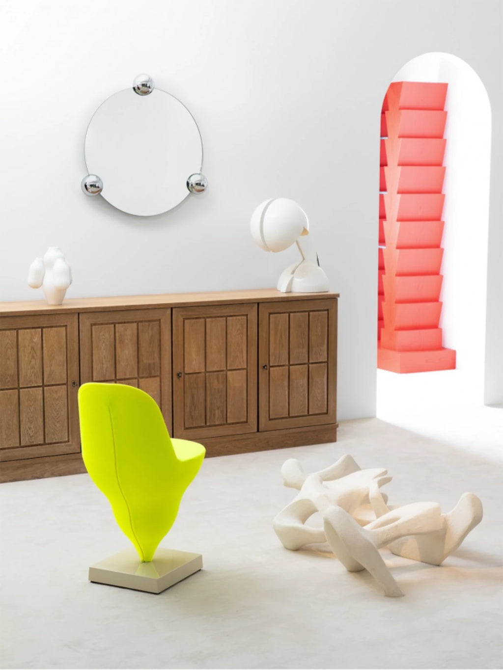 Moustache Sphera Orbit Mirror, a sleek circular mirror with three prominent chrome spheres, mounted above a wooden sideboard in a bright, contemporary room with a vibrant yellow chair and abstract sculpture.