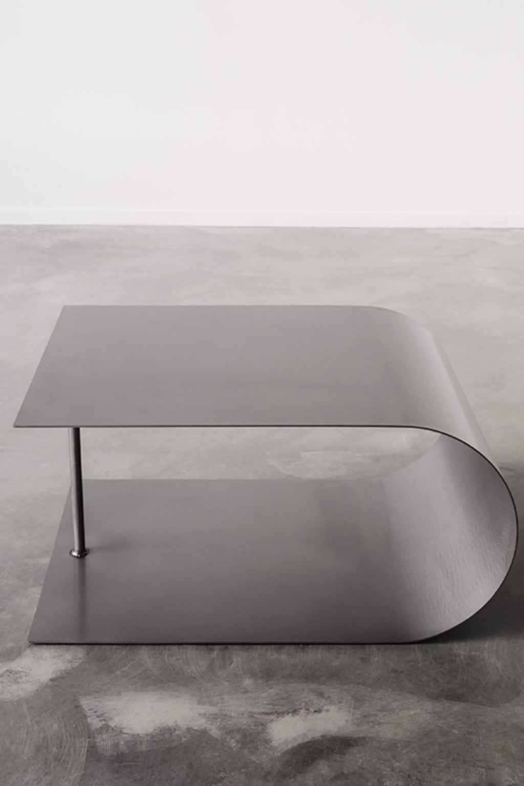 NG DESIGN Object 054 Coffee Table, a minimalist dark grey metal bent sheet coffee table with a single polished silver leg, displayed on a concrete floor. Perfect for modern living spaces.
