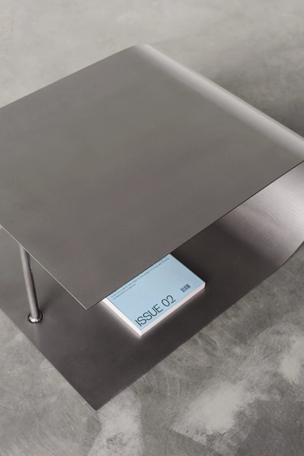 NG DESIGN Object 054 Coffee Table in a lifestyle shot. Features a dark gray, matte metal design with a curved upper and lower shelf, connected by a single metal leg. A light blue magazine labeled "ISSUE 02" rests on the lower shelf, showcasing its unique storage. This luxury minimalist coffee table is perfect for modern living spaces.
