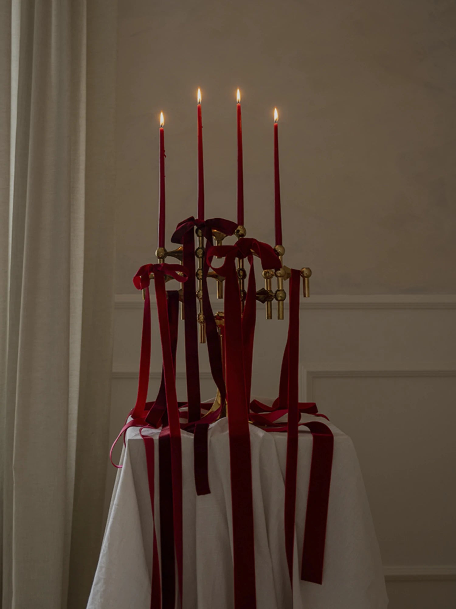 Decorative stoff nagel candle holder with red taper candles and red ribbons on a white tablecloth against a beige wall.