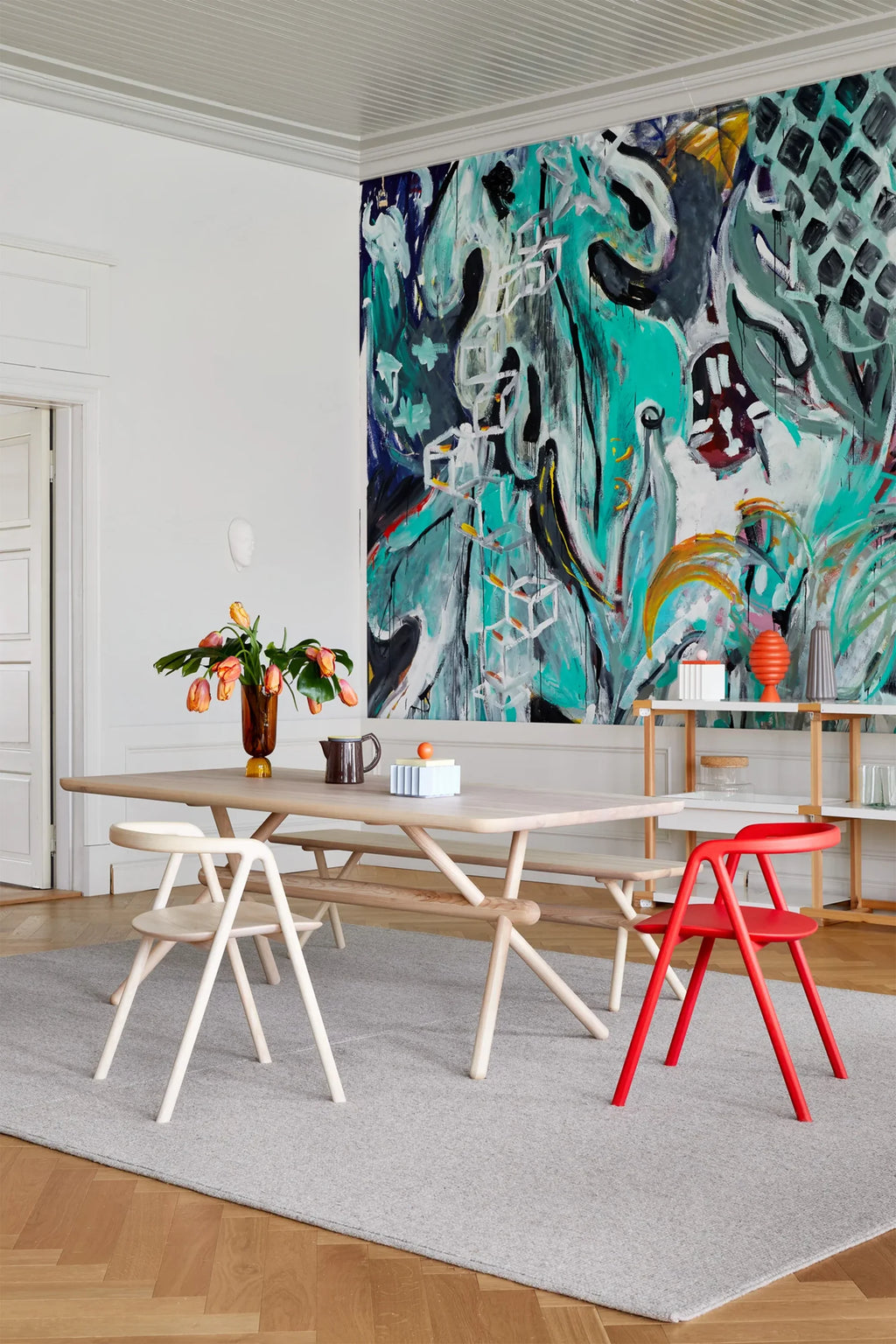 Made by Choice Laakso Dining Chair, a natural wood and a red chair, flanking a long wooden dining table in a spacious room with a large, vibrant abstract wall art, creating a contemporary and inviting dining area.