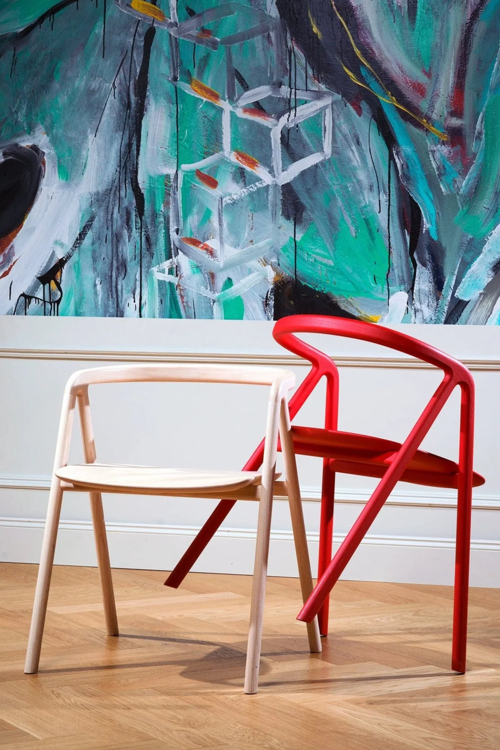 Made by Choice Laakso Dining Chair, two chairs, one natural wood and one vibrant red, positioned against a wall with a large, colorful abstract painting, showcasing their modern and versatile design in a stylish interior.