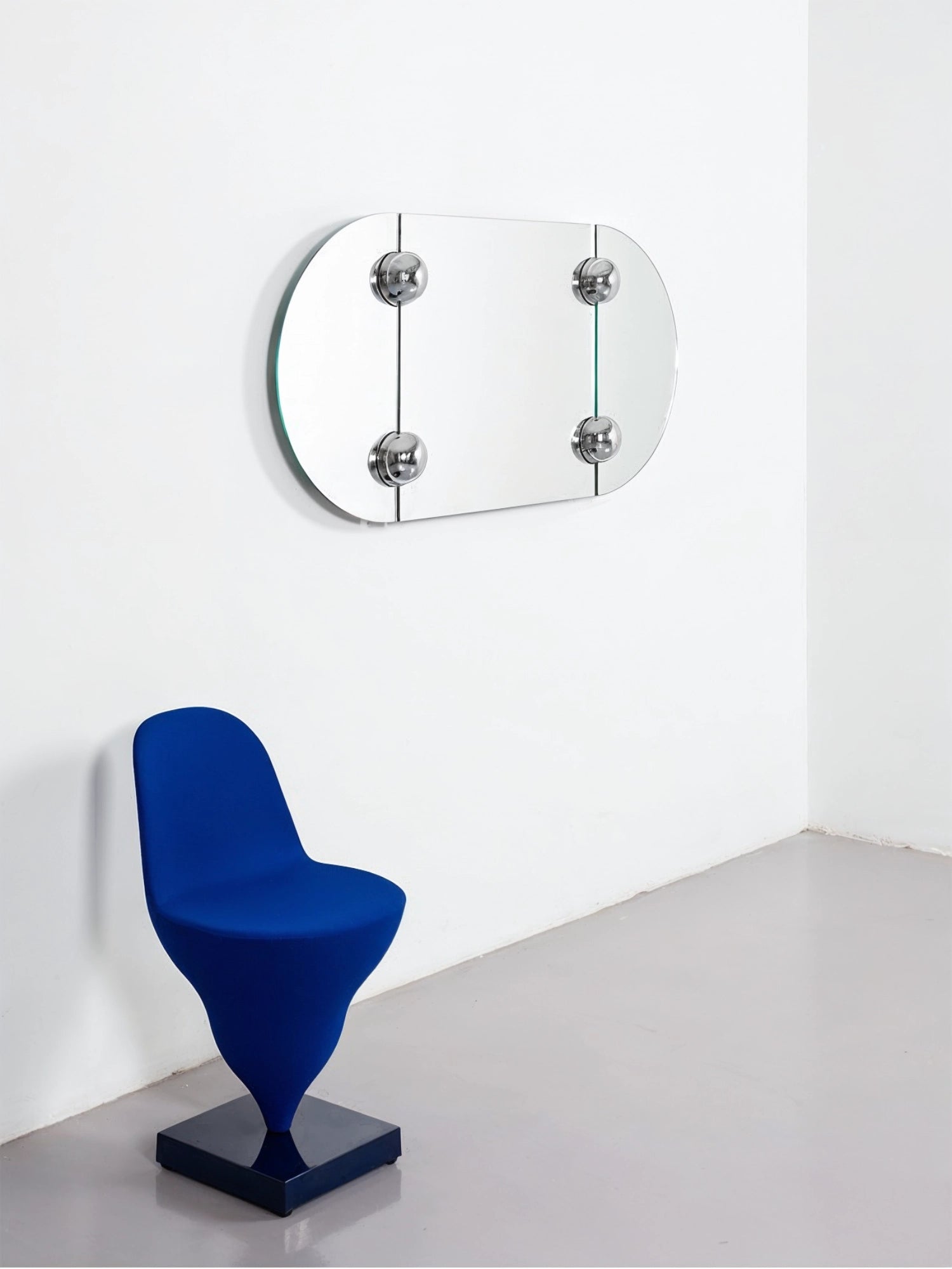 Moustache Sphera Oblong Mirror, a stylish lifestyle image showcasing the modern, elongated oval mirror with chrome spherical fasteners, mounted on a white wall above a vibrant blue sculptural chair, adding a touch of contemporary elegance to the room.