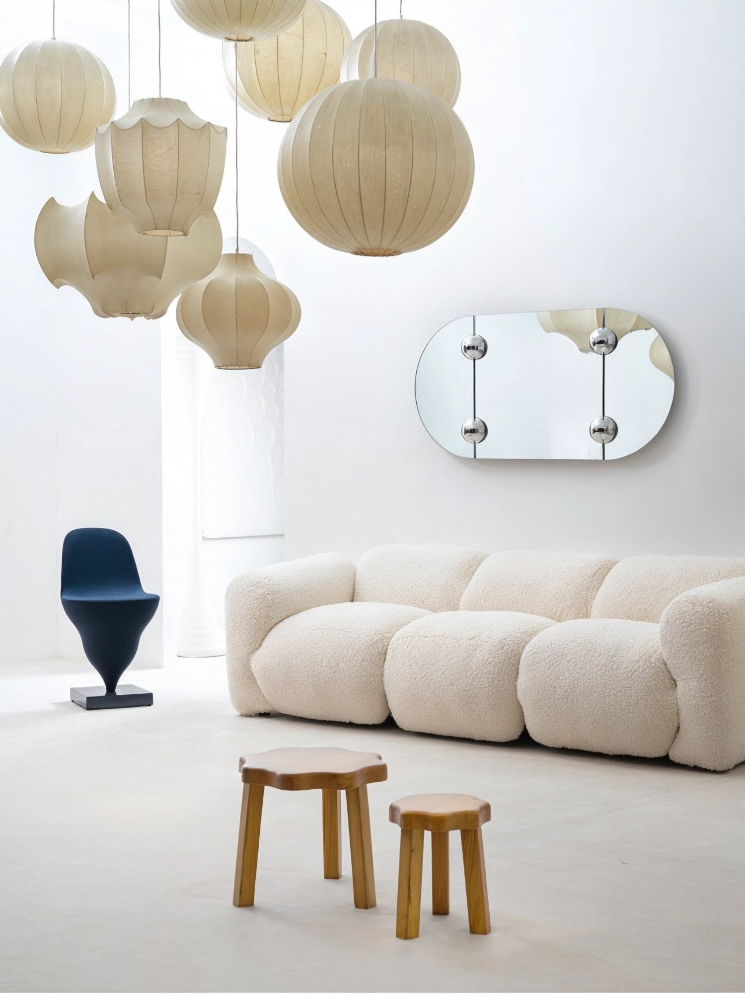 Moustache Sphera Oblong Mirror, a sophisticated lifestyle shot featuring the elongated oval mirror with its distinctive chrome spherical fasteners, reflecting a collection of elegant pendant lights, positioned above a cream bouclé sofa in a bright, modern living space.