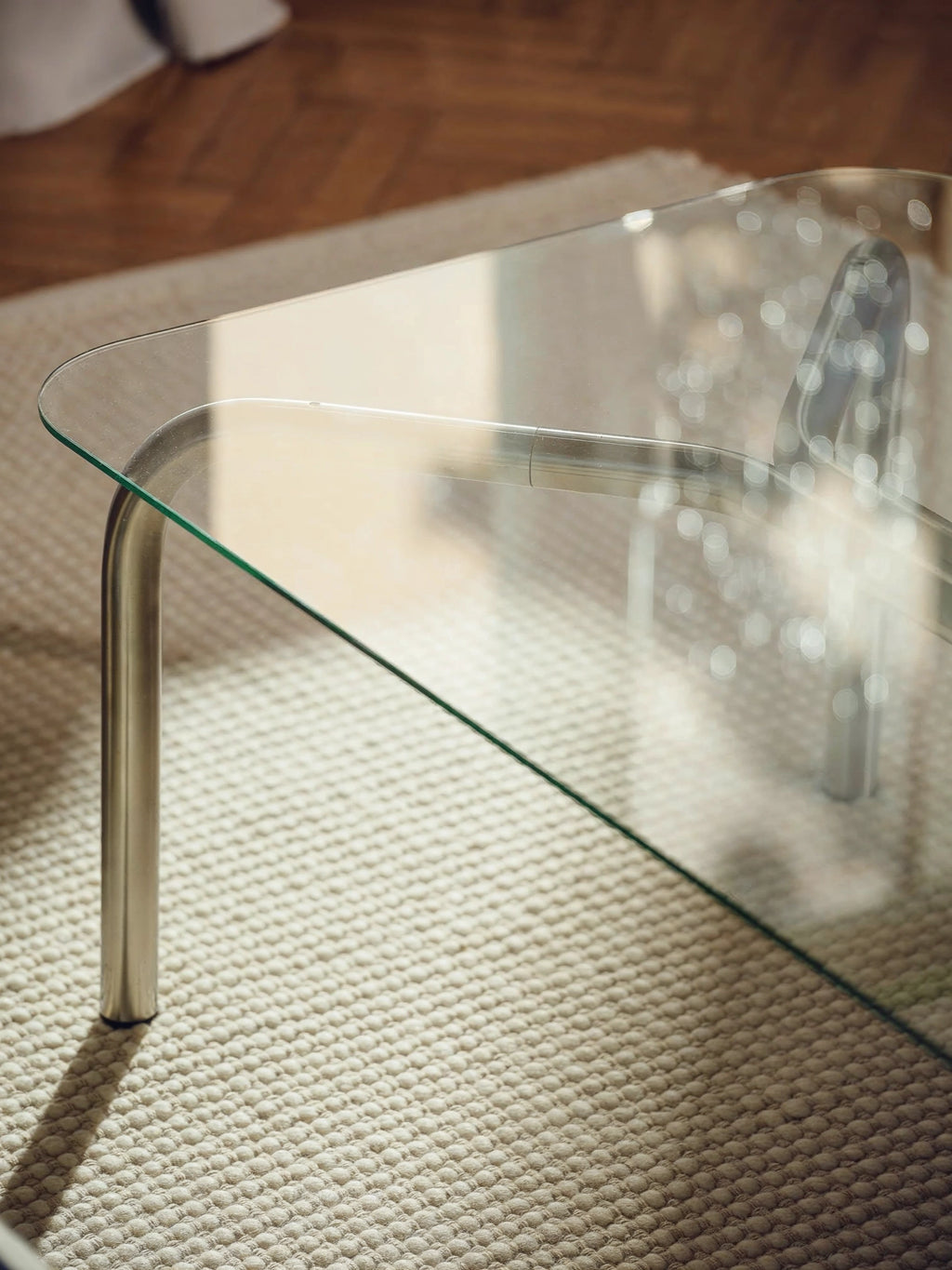 noo.ma Uku Coffee Table, a detailed shot focusing on the clear glass tabletop and one of the curved silver metal legs. The transparent surface reflects ambient light, showcasing the clean lines and modern design, resting on a textured cream rug.