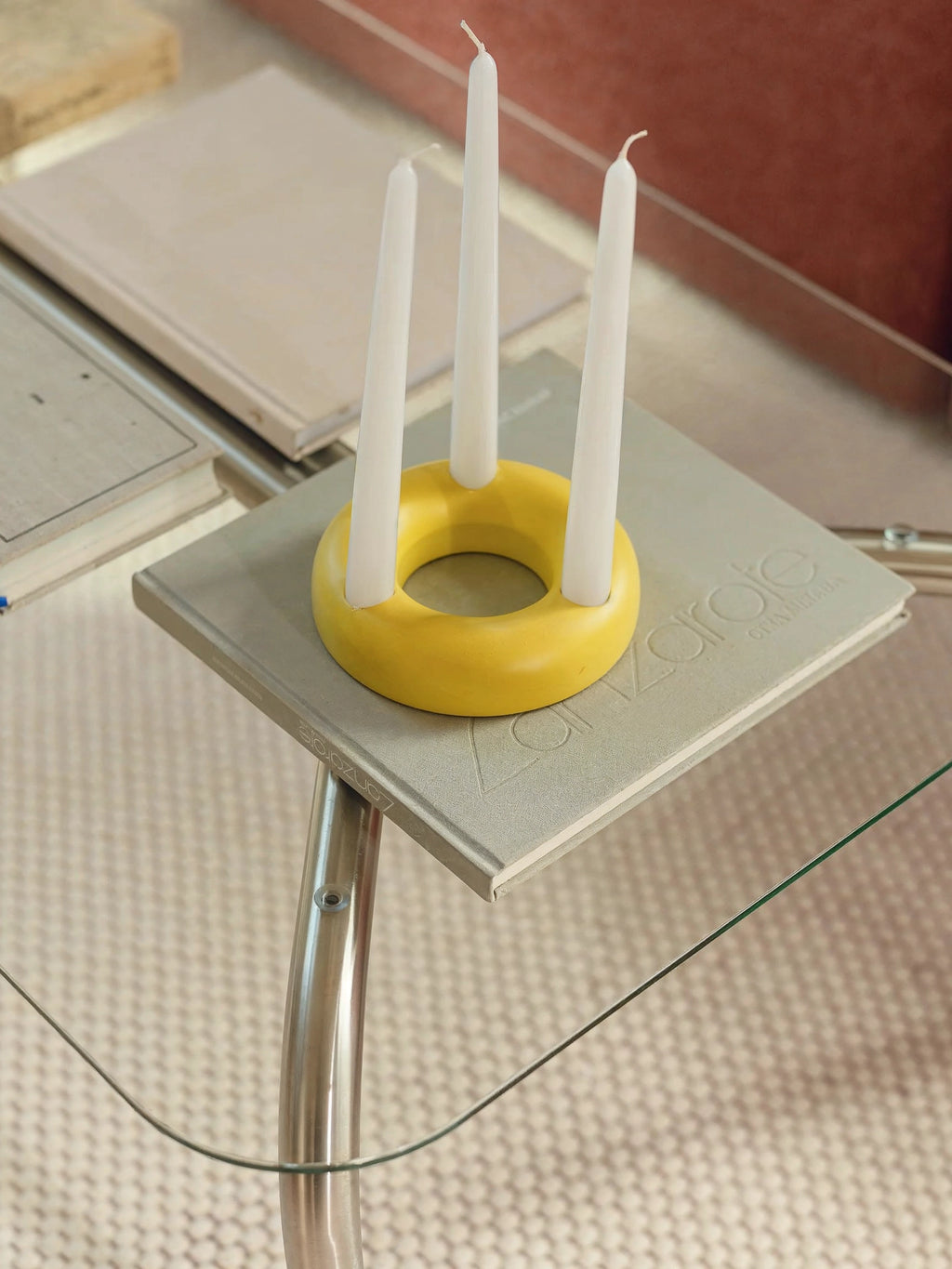 noo.ma Uku Coffee Table, a close-up of the rectangular glass surface, adorned with a light gray book and a vibrant yellow circular candle holder featuring three white taper candles. The modern design and functional styling are clearly visible.