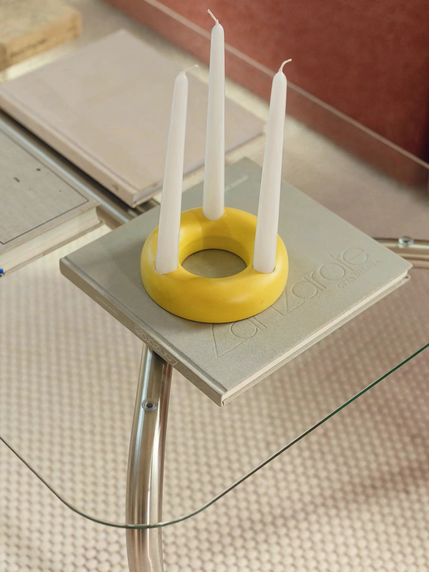 noo.ma Uku Coffee Table, a close-up of the rectangular glass surface, adorned with a light gray book and a vibrant yellow circular candle holder featuring three white taper candles. The modern design and functional styling are clearly visible.